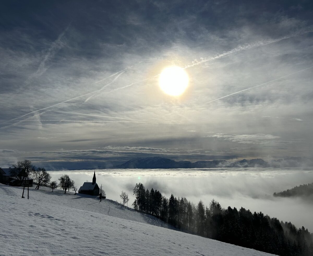 A winter landscape view showing a snow-covered hill with a church, a fog-filled valley, and distant mountains under a bright sun.