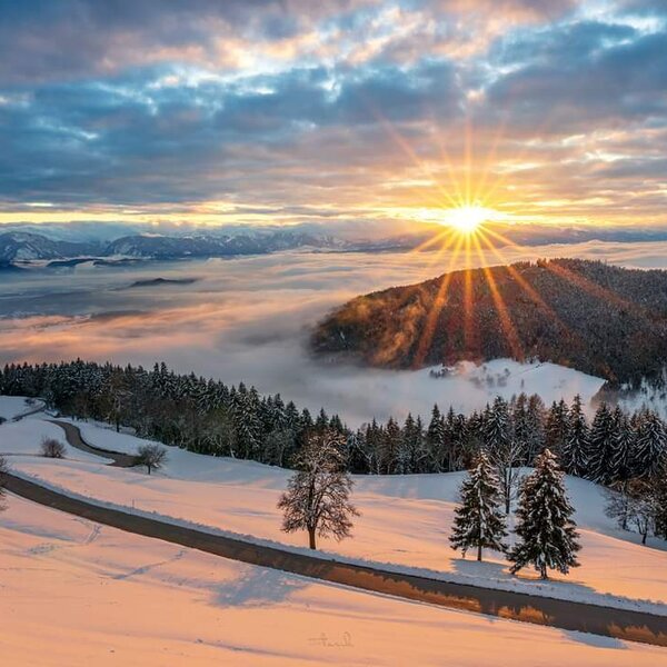 The winter landscape near the Farm House features snow-covered hills, pine forests, and a winding road, with a bright sunrise over a cloud-filled valley and distant mountains.