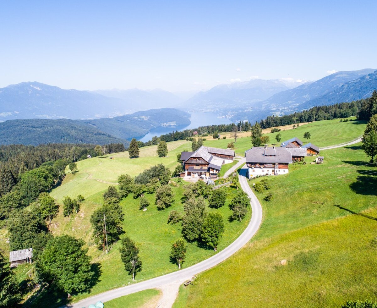 The farm house features multiple buildings set on a green hillside, with a winding road and views of a lake and mountains.