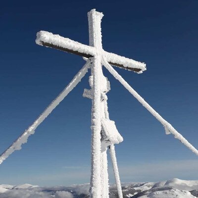 Snow-covered summit cross under a blue sky with views of the surrounding snowy mountains, typical of the winter landscape near the farmhouse.