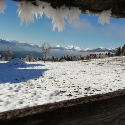 Winter view from the farmhouse with hoarfrost on the wooden frame, snow-covered fields, and mountains in the background.