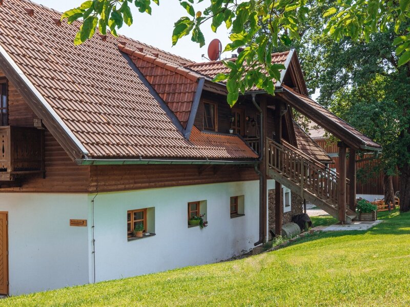 The exterior of the farm house features a tiled roof, wooden upper floor with a balcony, an external wooden staircase, and a surrounding green garden.