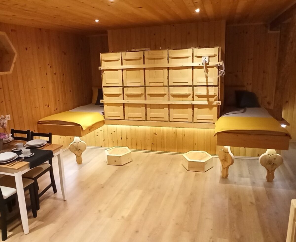 The wood-paneled room in the farm house features a dining table for four, two elevated beds, a prominent wall of wooden compartments, and decorative hexagonal wall shelves.
