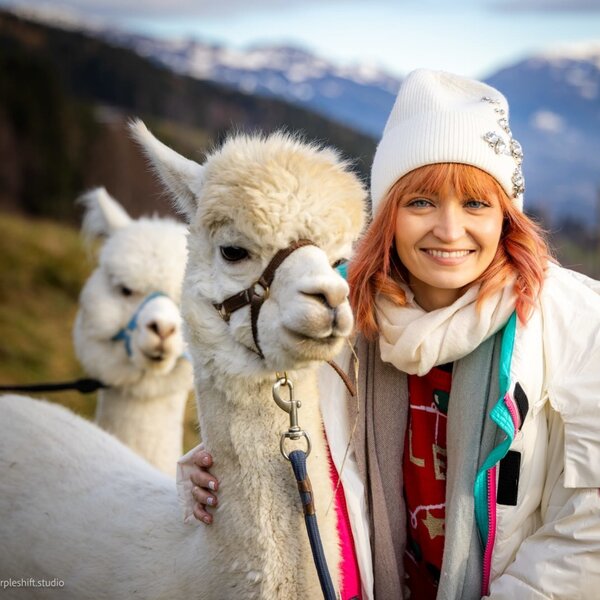 A smiling woman with an alpaca at the farmhouse, surrounded by mountains.