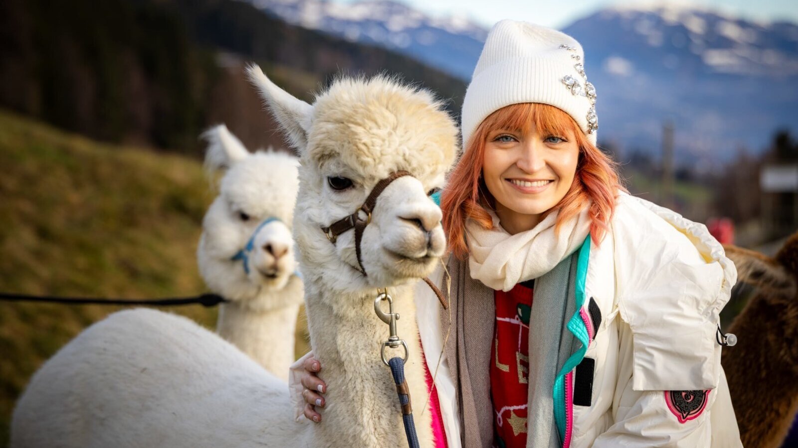 A smiling woman with an alpaca at the farmhouse, surrounded by mountains.