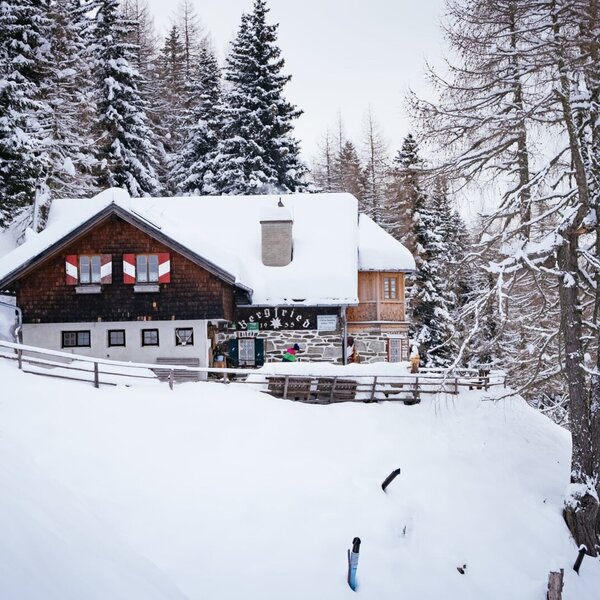 The Bergfried Alpine Hut stands covered in snow, nestled among snow-laden trees in a mountain setting.