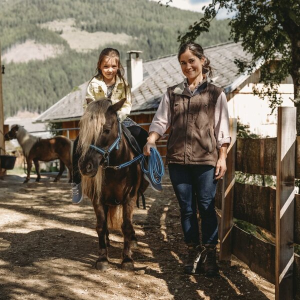 A child riding a pony led by an adult on the grounds of the farm house.