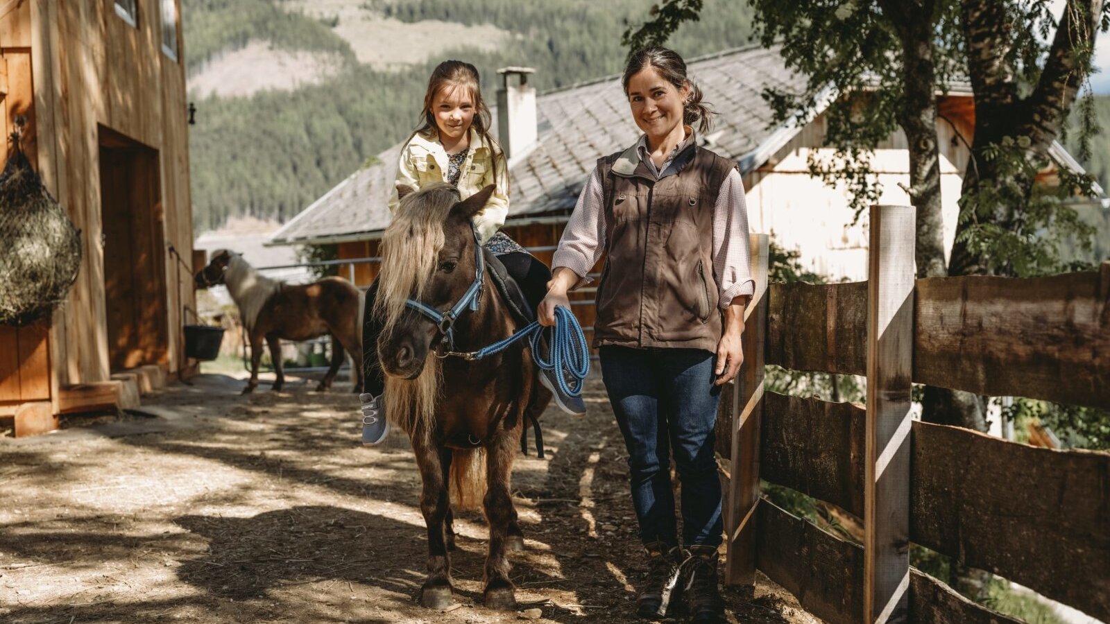 A child riding a pony led by an adult on the grounds of the farm house.