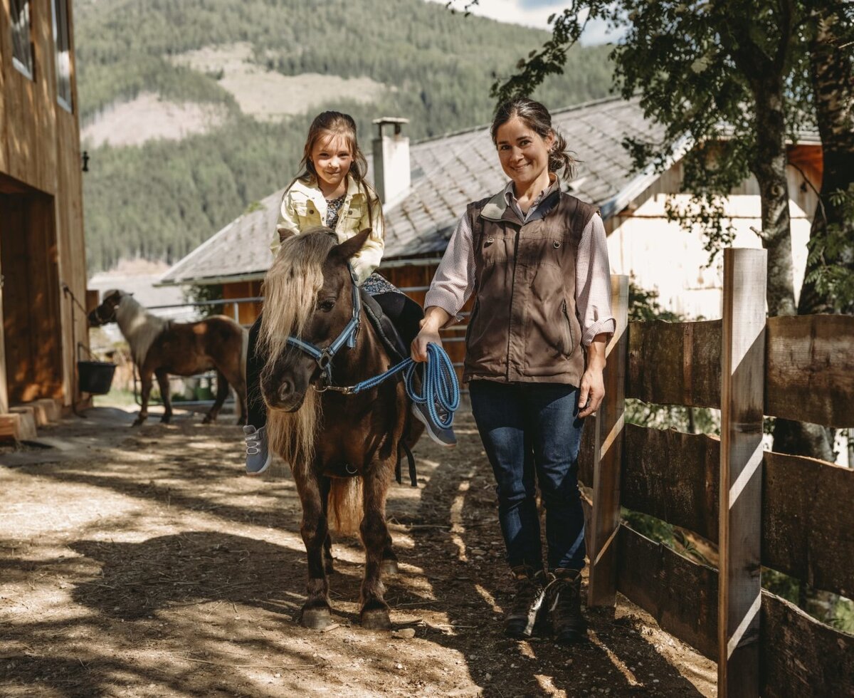 A child riding a pony led by an adult on the grounds of the farm house.