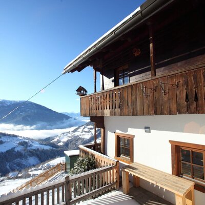 The farm house exterior features traditional wooden balconies and a snow-covered terrace with a wooden bench, offering views of the surrounding mountainous landscape.