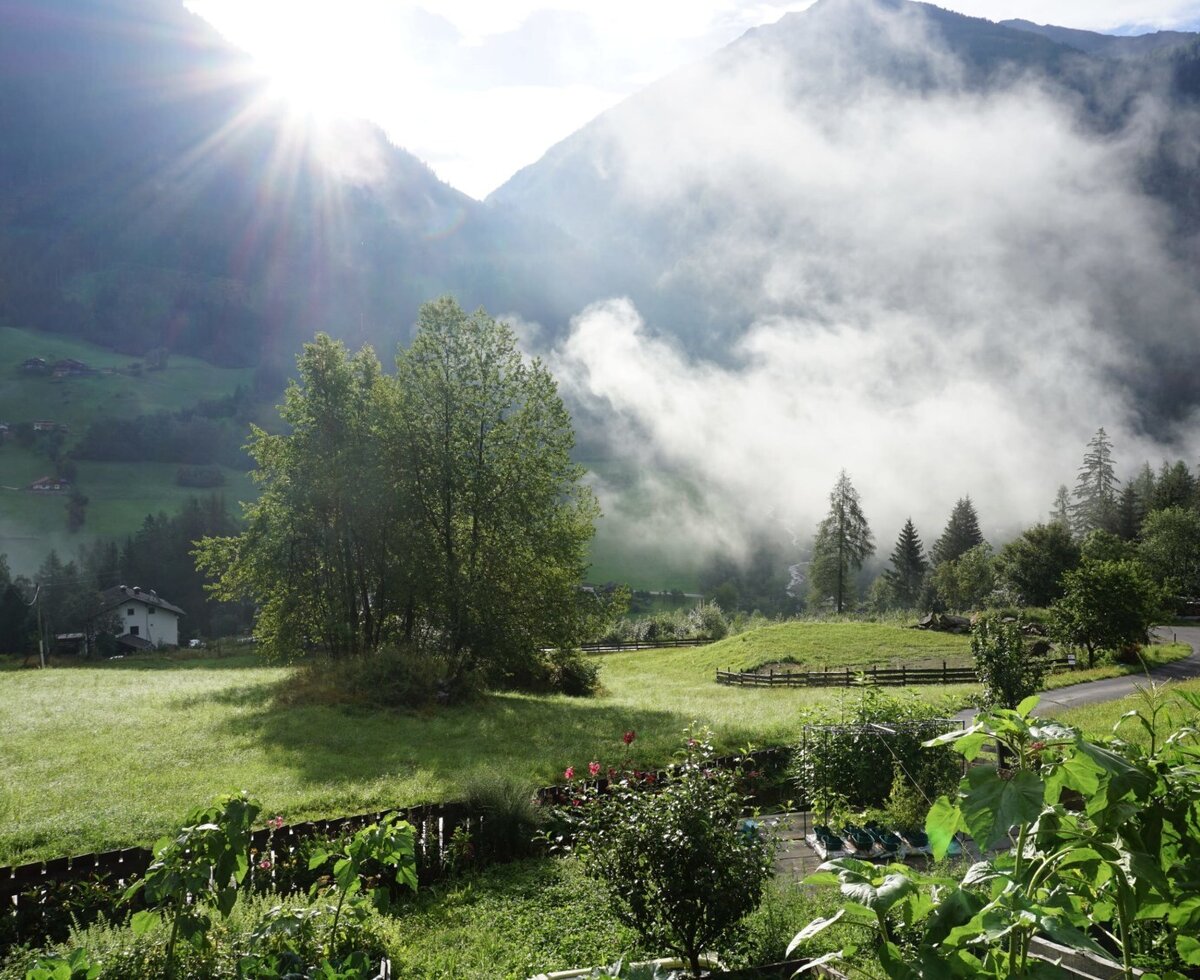 View of the Mölltal valley from the Farm House, featuring sunlit mountains, misty fields, and a foreground garden.