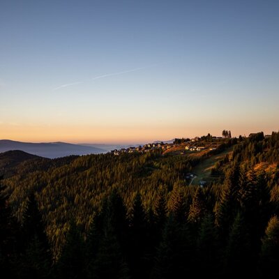 The Alm is situated amidst a forested mountain landscape with a view of a village on the hillside at sunset.