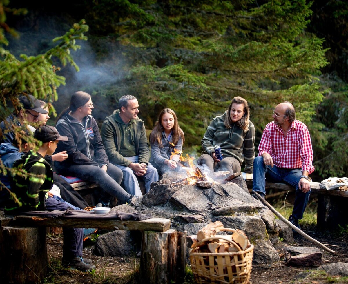 Guests of the Alm sitting around a campfire in the forest, with wooden benches and a basket of firewood.