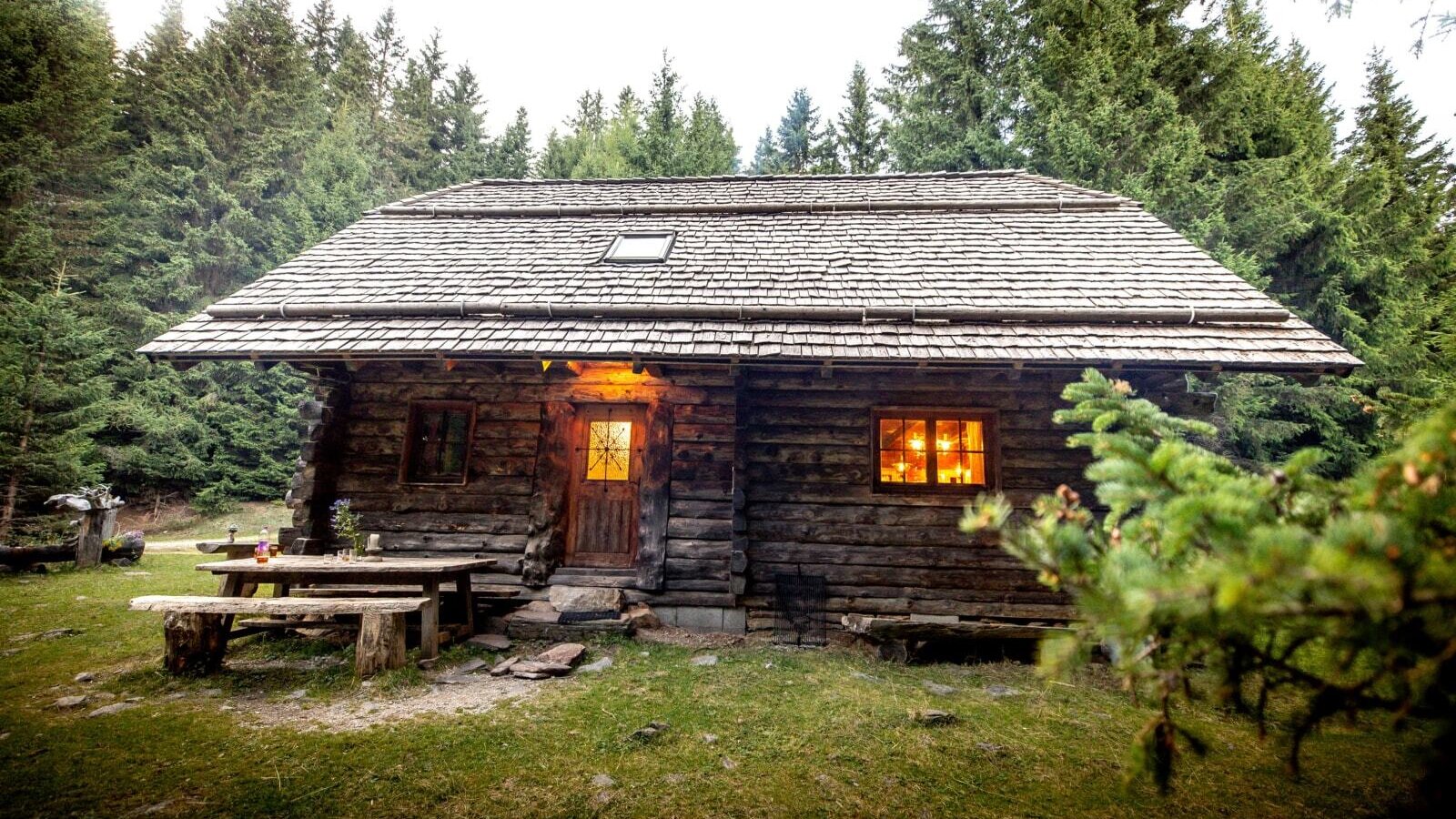 The exterior of the Alm, featuring wooden walls, a shingled roof, illuminated windows, and a wooden picnic table.