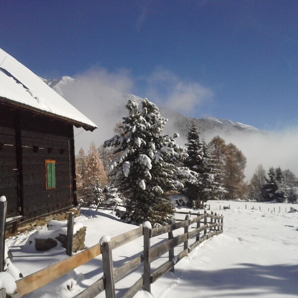 The Alpine Hut in a winter setting, showing its snow-covered exterior, wooden fence, and surrounding snowy mountains.
