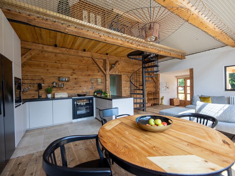 The open-plan living room in the farmhouse features a fully equipped kitchen, a wooden dining table, a sofa, a spiral staircase leading to the gallery, and a loft net.