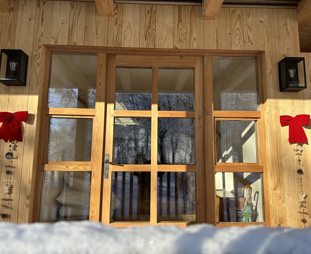 The farmhouse exterior with its large wooden-framed glass door and window unit, wall lights, and decorative accents, set against a snowy foreground.