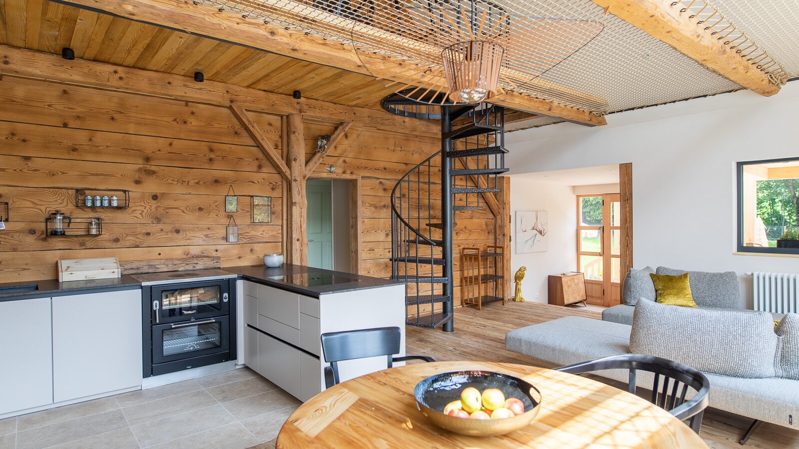 The open living space of the Ferienhaus features a fully equipped kitchen, dining table, sofa, and a spiral staircase leading to the gallery with a loft net.