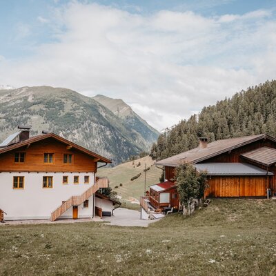 The farm house exterior features two traditional buildings with wooden details, set against a backdrop of mountains and forests.