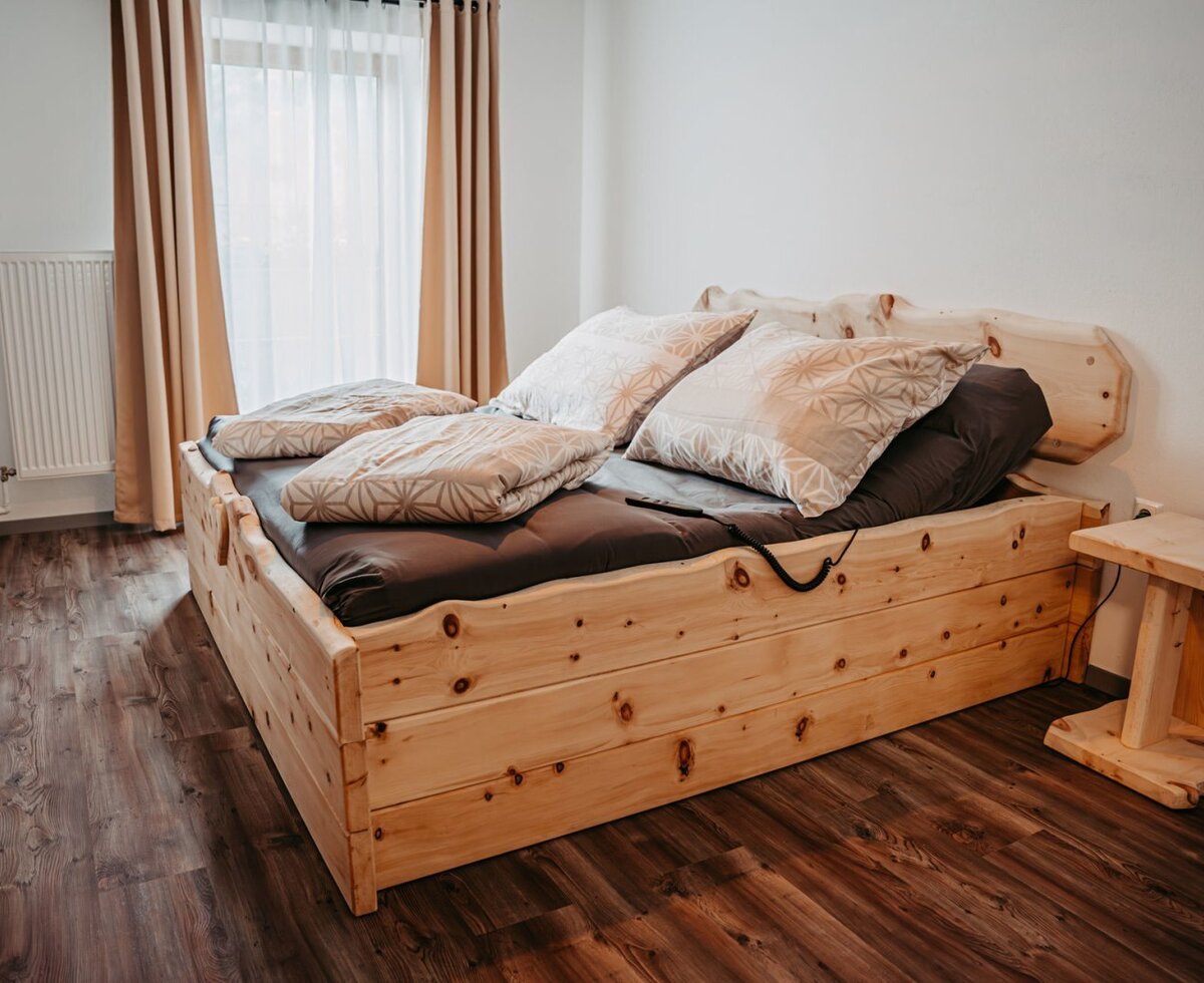 Bedroom in the farm house with a wooden adjustable bed, dark brown bedding, a wooden side table, and a window with sheer and opaque curtains.