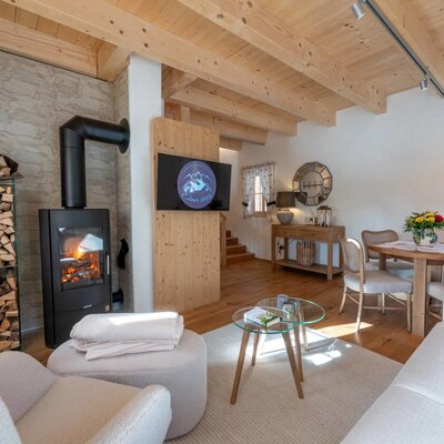 The traditional living room of the farmhouse, featuring a fireplace, stacked firewood, a dining table, and a sofa under a wooden beam ceiling.