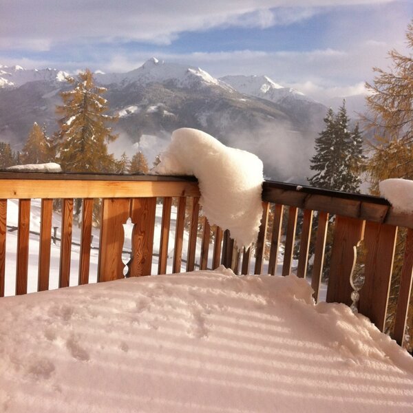 The snow-covered balcony of the Alpine Hut, featuring a wooden railing, provides views of the mountains and wooded valley.