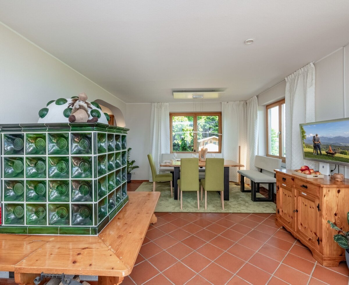 The living area of the farmhouse, featuring a green tiled stove, a dining table, and a wall-mounted TV.