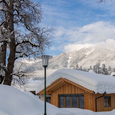 The Farm House in a winter setting, with snow covering its wooden exterior and the surrounding mountainous landscape.