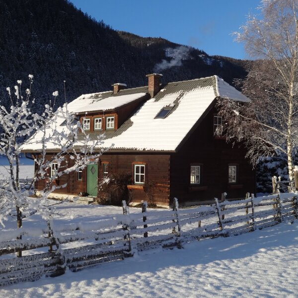 The exterior of the wooden Alpine Hut, featuring a snow-covered roof, a green entrance door, and a snowy fence in a mountain landscape.