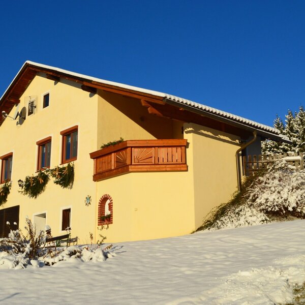 The yellow exterior of the Farm House features wooden window frames and a balcony, set against a snowy landscape with evergreen trees.