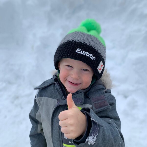 A child in a winter jacket and hat gives a thumbs-up against a snowy background, suggesting winter activities for families at the farm house.