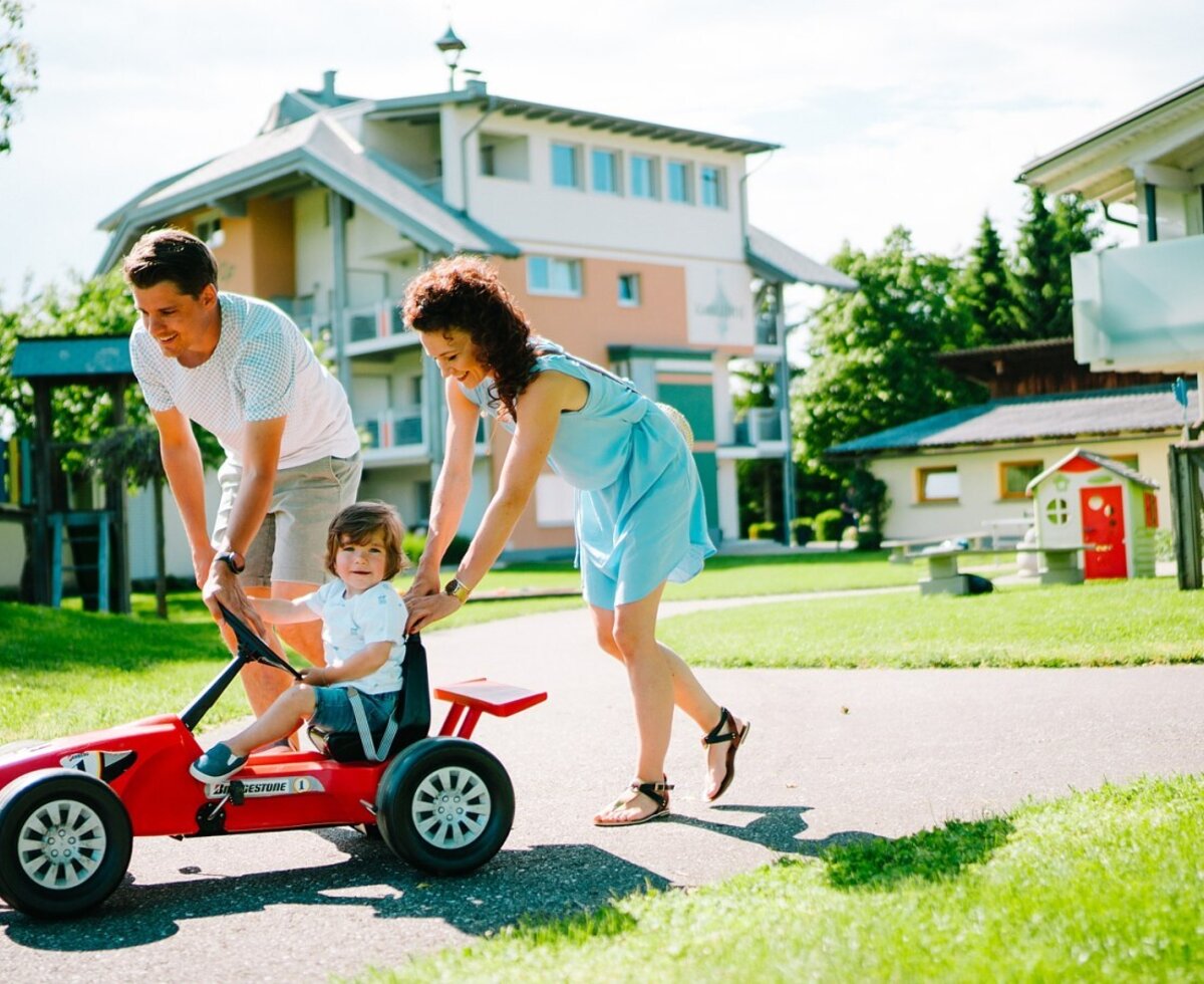 A family plays with a red pedal car on the paved path in the garden of the farmhouse, with a children's play area in the background.