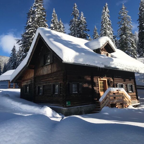 The wooden Alpine Hut exterior, featuring a snow-covered roof and surrounded by snowy landscape and pine trees.