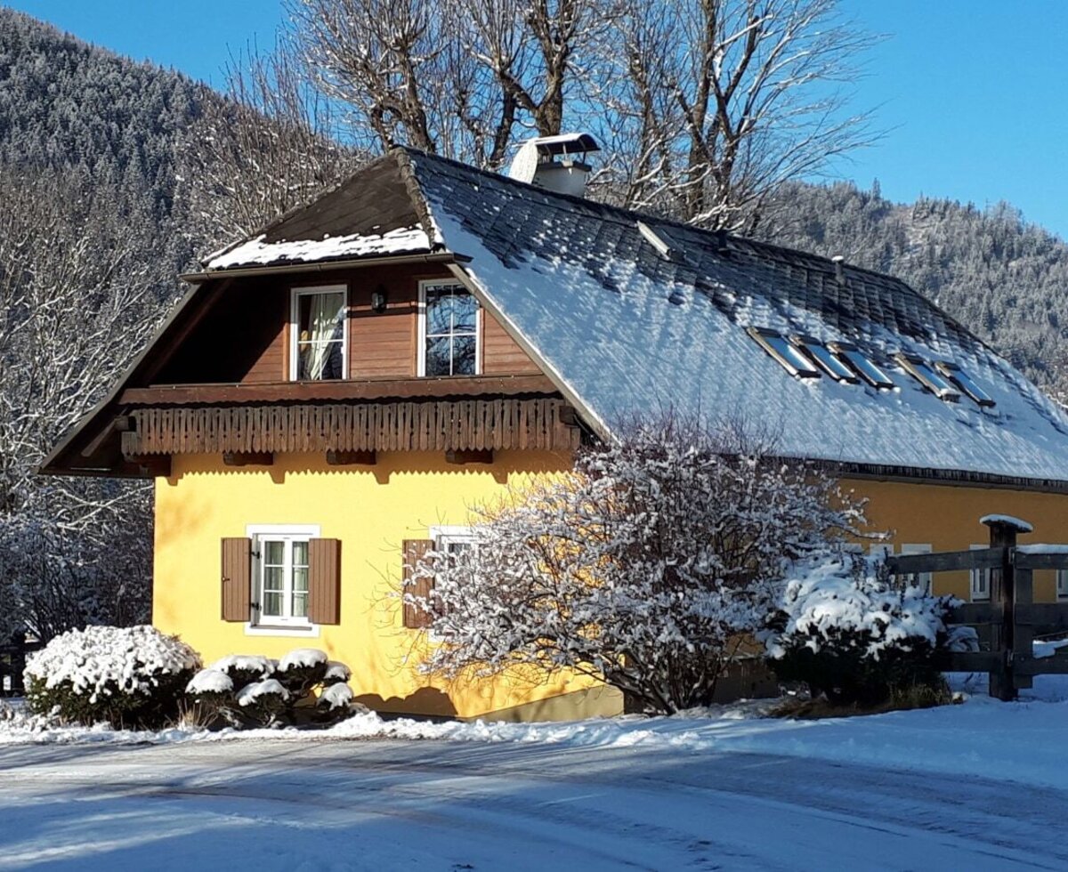 The yellow Farm House features a snow-covered roof and surrounding grounds, set against a backdrop of snow-dusted mountains and trees.
