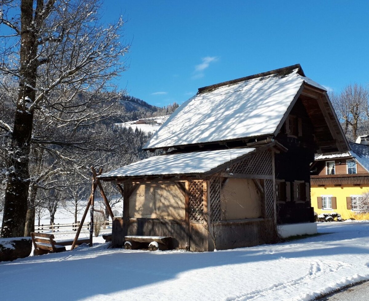 The Farm House exterior during winter, showcasing snow-covered wooden buildings, an outdoor seating area, and forested mountains in the background.