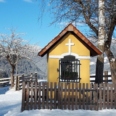 A small yellow wayside shrine with a cross, enclosed by a wooden fence in the snowy landscape surrounding the Farm House.