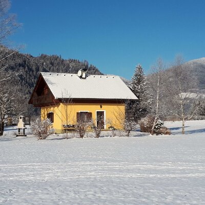 The yellow Farm House, with its snow-covered roof and balcony, stands amidst snowy fields and mountains.