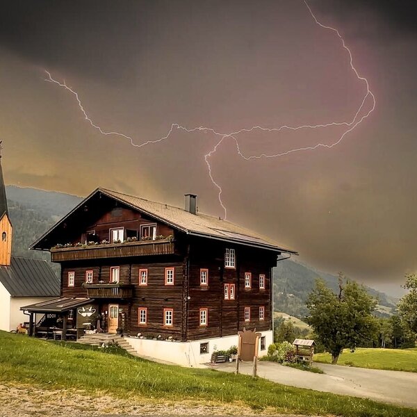 A traditional wooden farmhouse and a small chapel set against a dramatic stormy sky over the surrounding mountains.