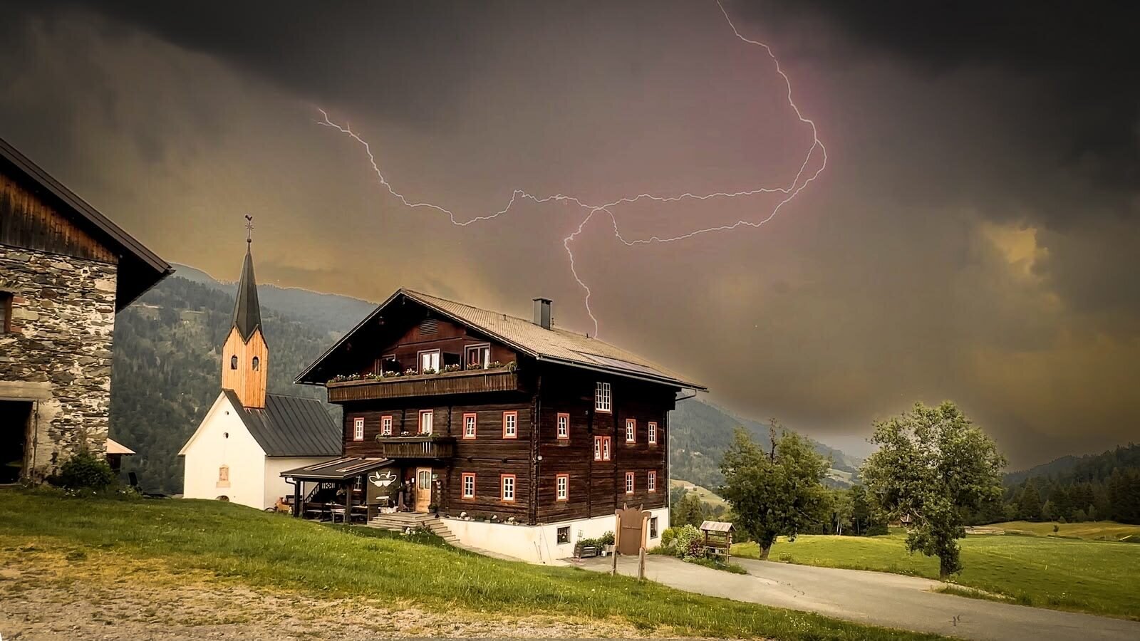 A traditional wooden farmhouse and a small chapel set against a dramatic stormy sky over the surrounding mountains.