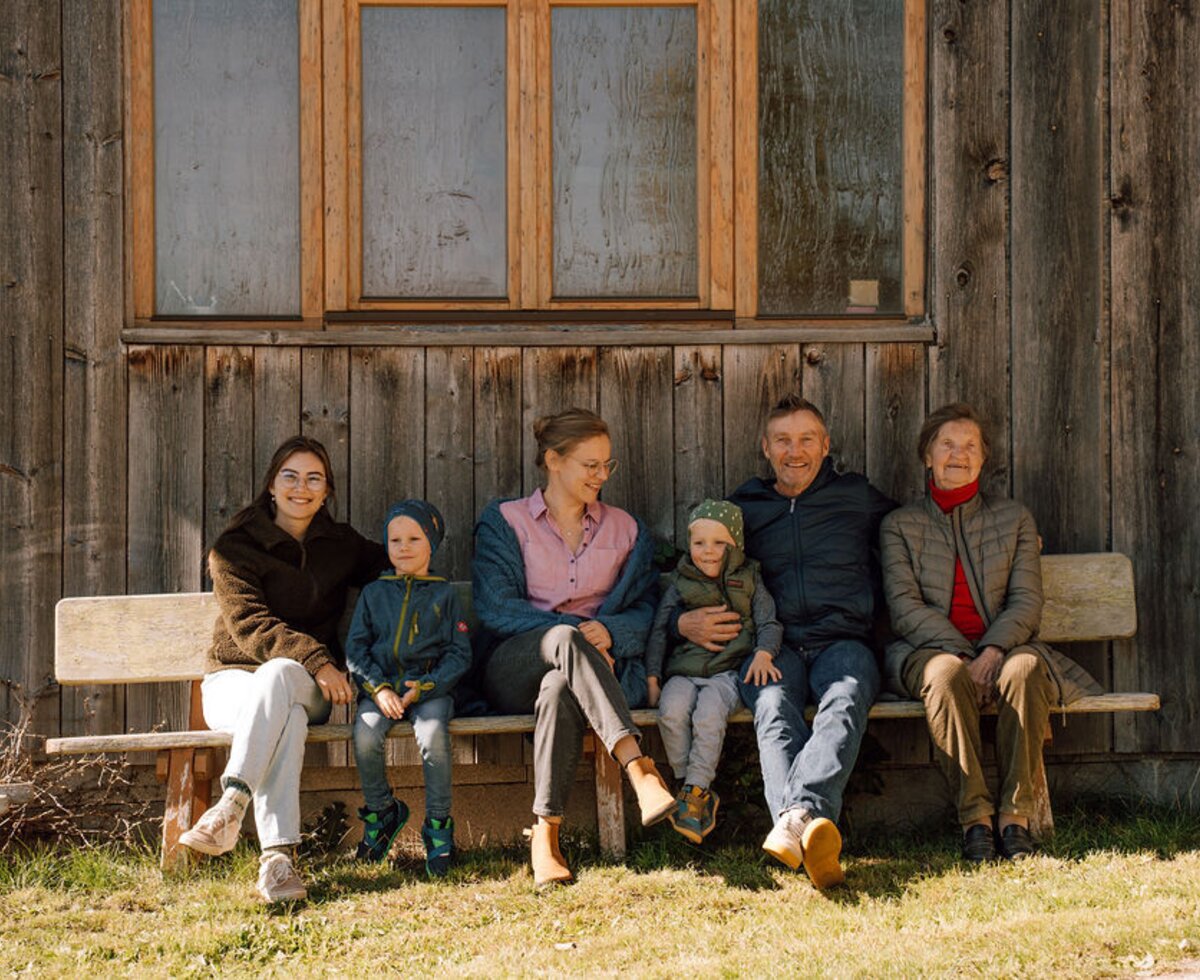 The farmhouse family sitting on a wooden bench in front of the wooden facade.
