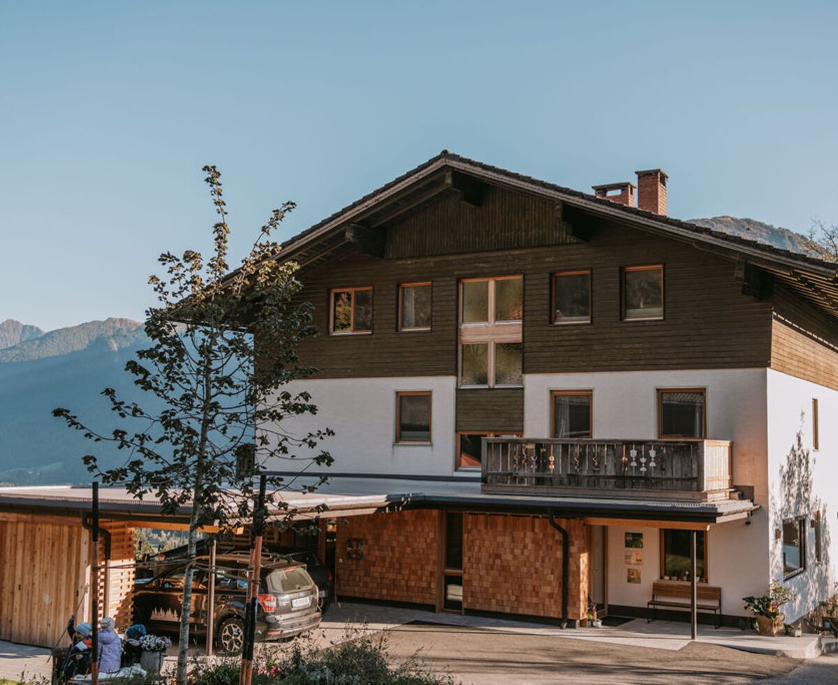 The exterior of the farmhouse, featuring traditional wood and plaster facade, a carport, and mountain views.