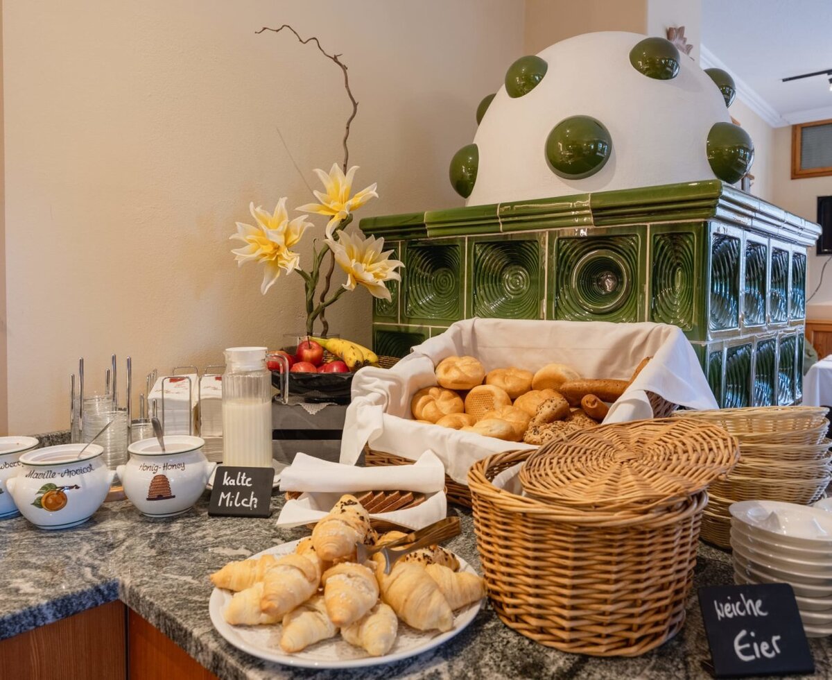 Breakfast buffet at the farmhouse featuring fresh bread rolls, croissants, jam, honey, milk, and fruit, alongside a traditional tiled stove.