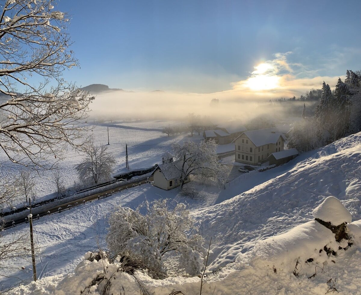 The snow-covered Gschlosserhof farmhouse in an idyllic winter landscape with gentle sunshine and misty fog.