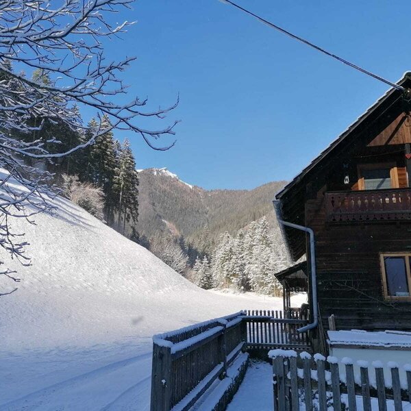 The farm house exterior featuring a wooden balcony and a fence, surrounded by a snow-covered landscape with mountains and evergreen trees.
