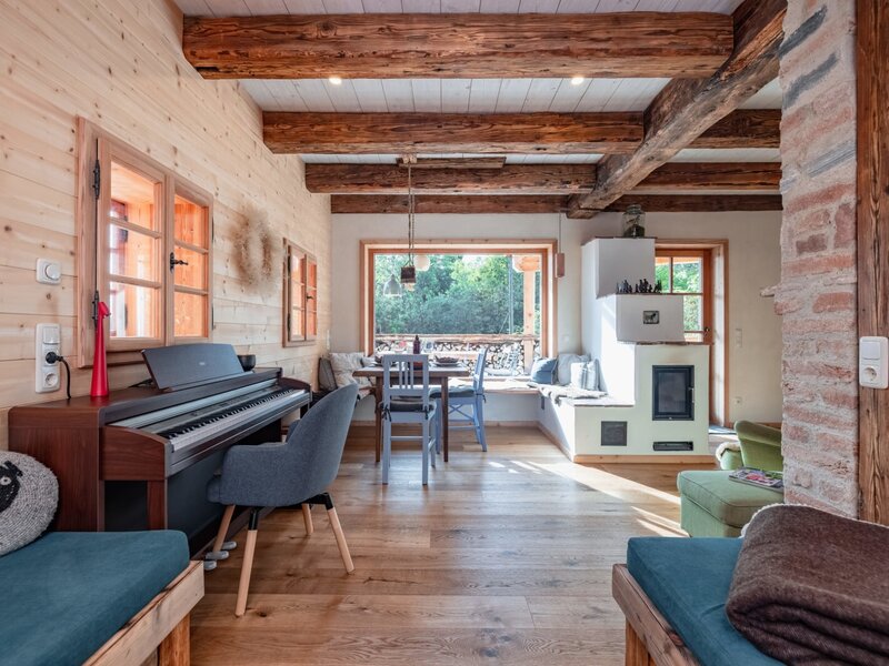 The living area of the holiday home, featuring a piano, a dining table by the window, a traditional tiled stove, and exposed wooden beams.
