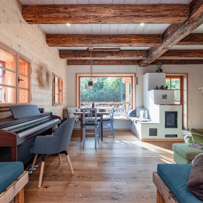The living area of the holiday home, featuring a piano, a dining table by the window, a traditional tiled stove, and exposed wooden beams.