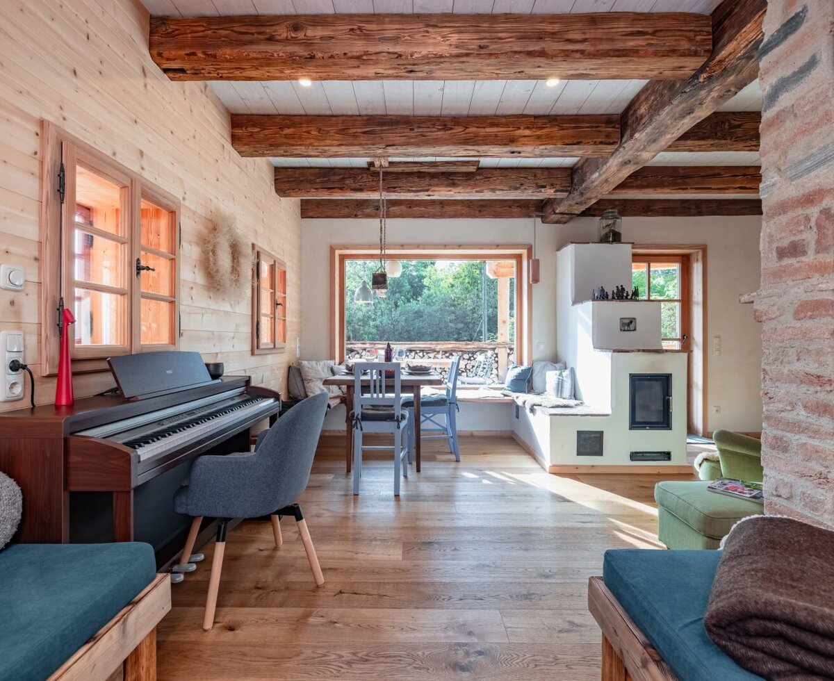 The living area of the holiday home, featuring a piano, a dining table by the window, a traditional tiled stove, and exposed wooden beams.