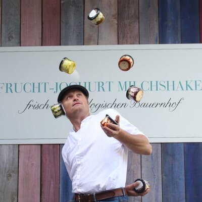 A man juggling yogurt cups in front of a sign advertising fruit yogurt milkshakes from the organic farm of the Ferienhaus.
