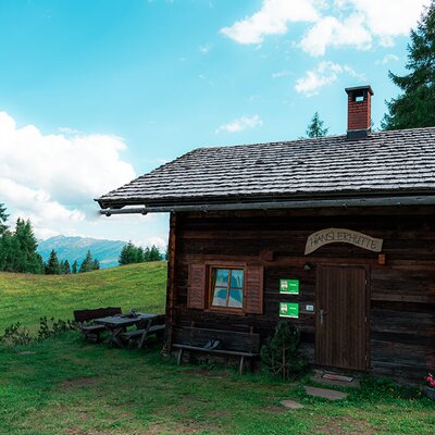 The Alpine Hut "Hanslerhütte" features an outdoor wooden picnic table and benches, set amidst alpine pastures with mountain views.