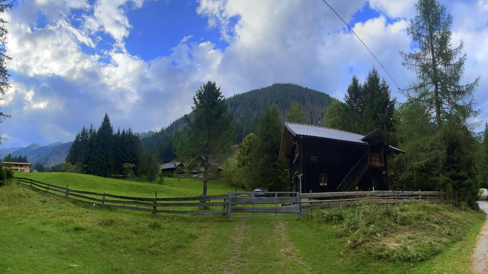 A rustic Alm set amidst lush green meadows, surrounded by dense pine trees and rolling mountain slopes.